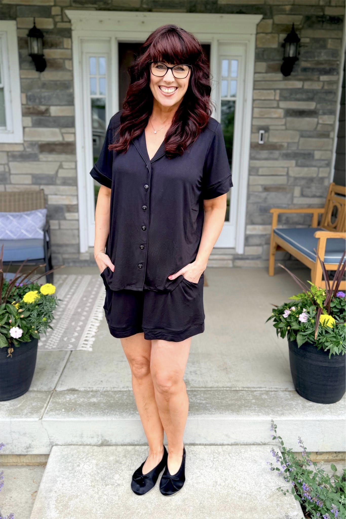 Woman modeling the LuLaRoe Candy PJ Set in Solid Black featuring a button-front short sleeve pajama top with contrast piping and matching relaxed-fit shorts with pockets in a classic black sleepwear set standing on a porch with plants and furniture.