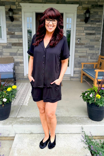 Woman modeling the LuLaRoe Candy PJ Set in Solid Black featuring a button-front short sleeve pajama top with contrast piping and matching relaxed-fit shorts with pockets in a classic black sleepwear set standing on a porch with plants and furniture.