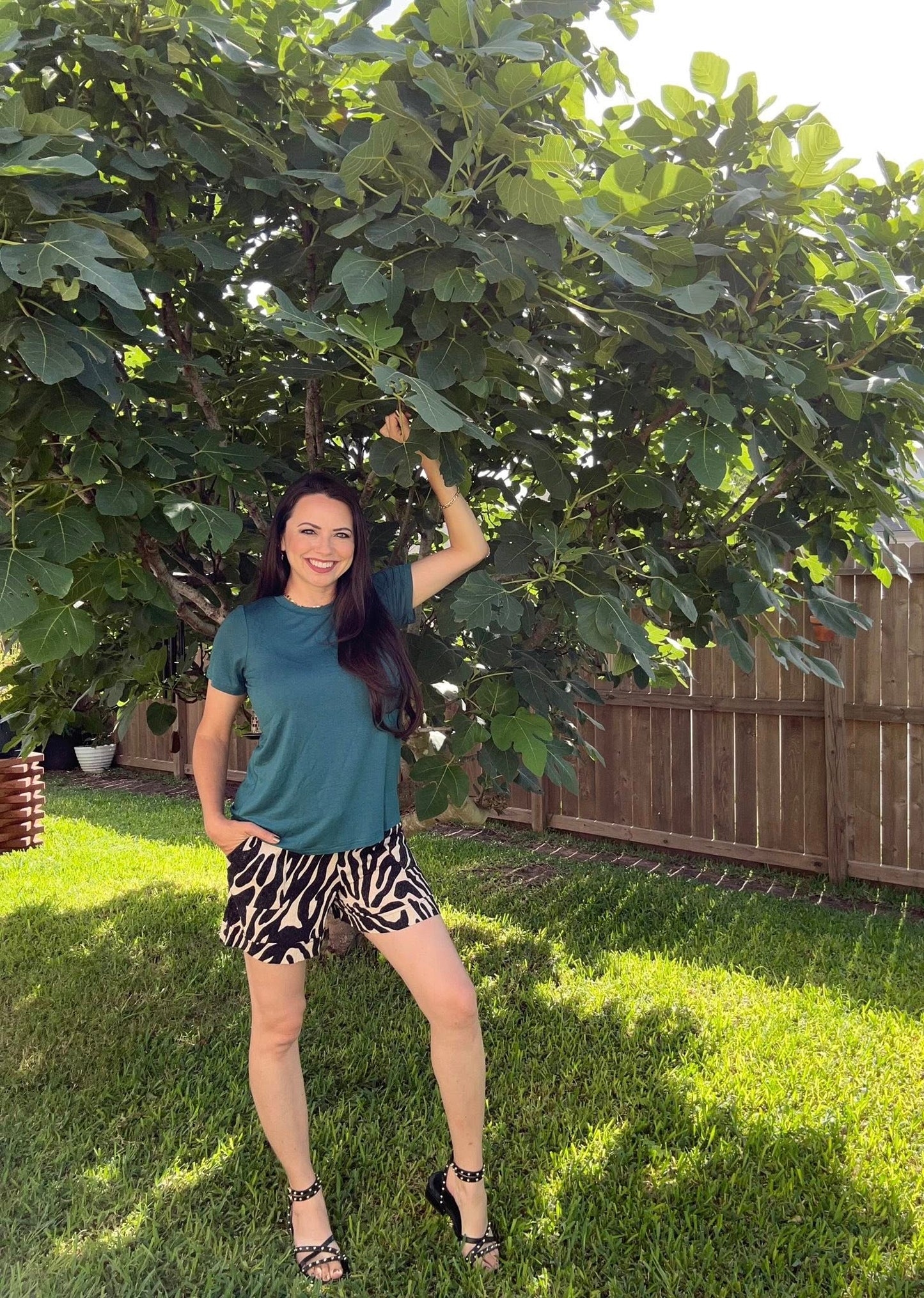 Woman wearing LuLaRoe Essential Tee in Solid Deep Teal featuring a rich blue-green color, classic crew neckline, short sleeves, and a semi-relaxed fit in soft luxe modal fabric in a garden with large green leaves in the background