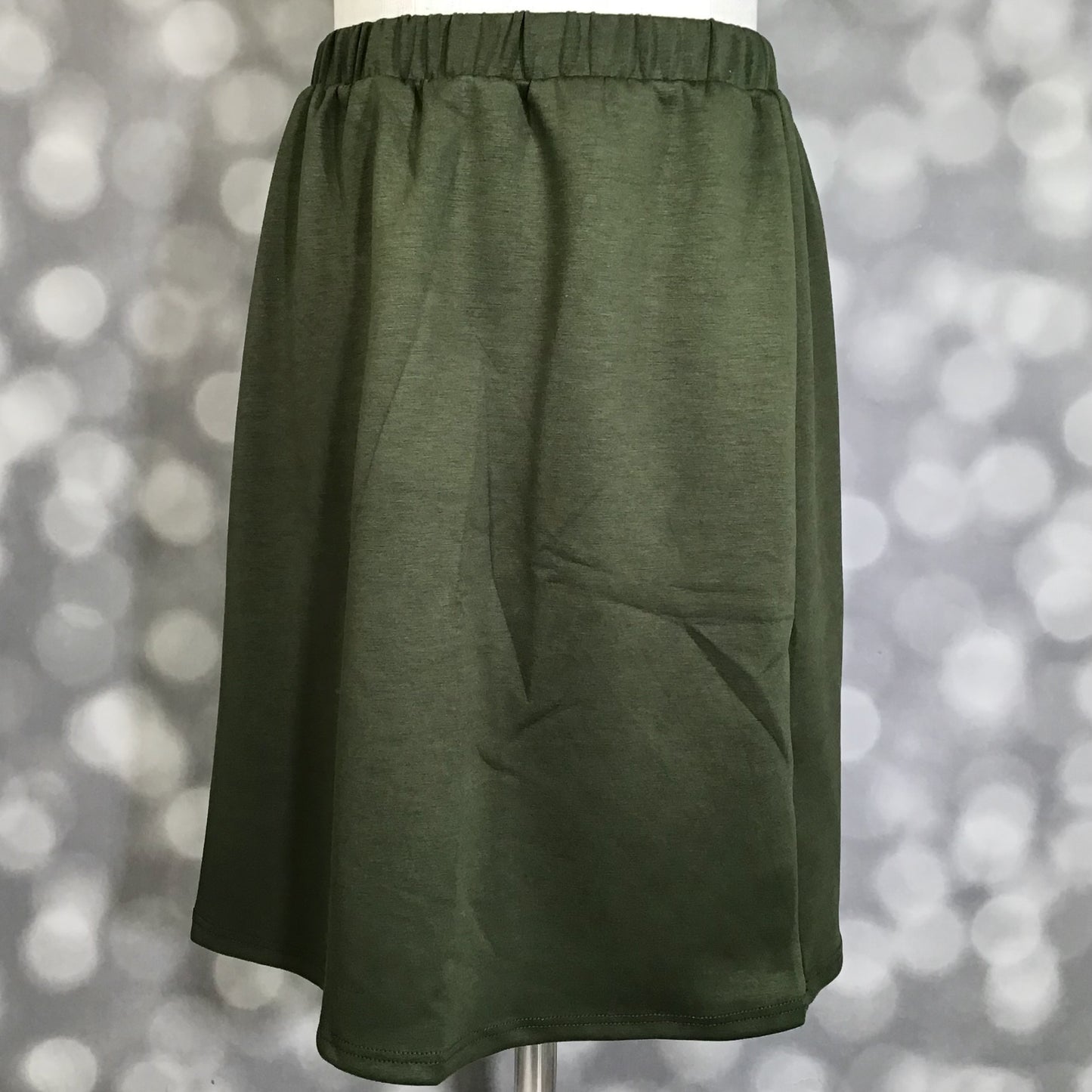 LuLaRoe Samantha Skirt in Solid Olive Grove, a women's pull-on midi skirt featuring a non-roll elastic waistband and soft Modal Scuba fabric. Deep olive green skirt with a flattering silhouette, ideal for casual outfits, fall fashion, workwear, and everyday comfort on a mannequin against a blurred background