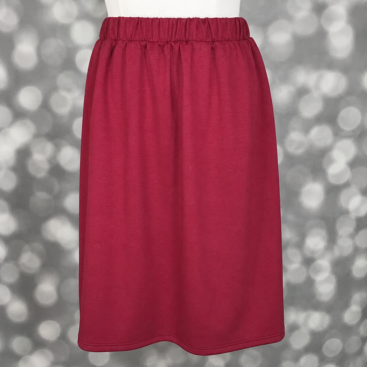 LuLaRoe Samantha Skirt in Solid Raspberry Wine, a women's pull-on midi skirt featuring a comfortable non-roll elastic waistband and soft Modal Scuba fabric. Rich raspberry red skirt with a flattering silhouette for everyday wear, work outfits, and seasonal styling on a mannequin against a blurred gray background
