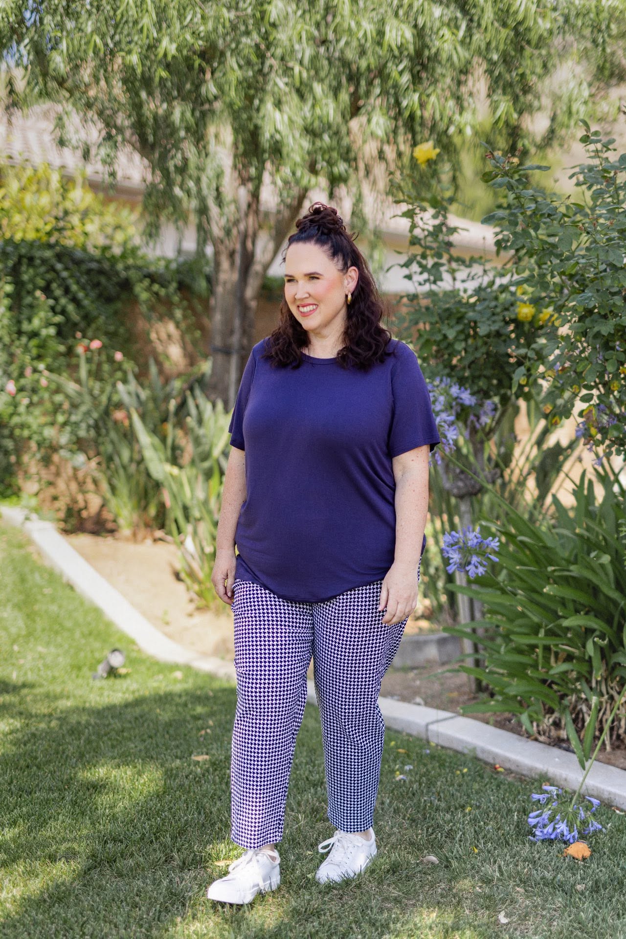 Woman wearing LuLaRoe Essential Tee in Solid Royal Iris featuring a rich blue-purple hue, classic crew neckline, short sleeves, and a semi-relaxed fit in soft luxe modal fabric standing outdoors in a garden wearing checkered pants.