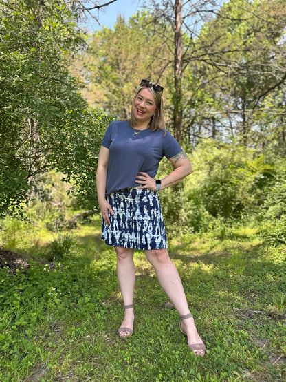 Woman standing in a forest wearing LuLaRoe Essential Tee in Solid Stormy Blue featuring a muted blue-gray color, classic crew neckline, short sleeves, and a semi-relaxed fit in soft luxe modal fabric.