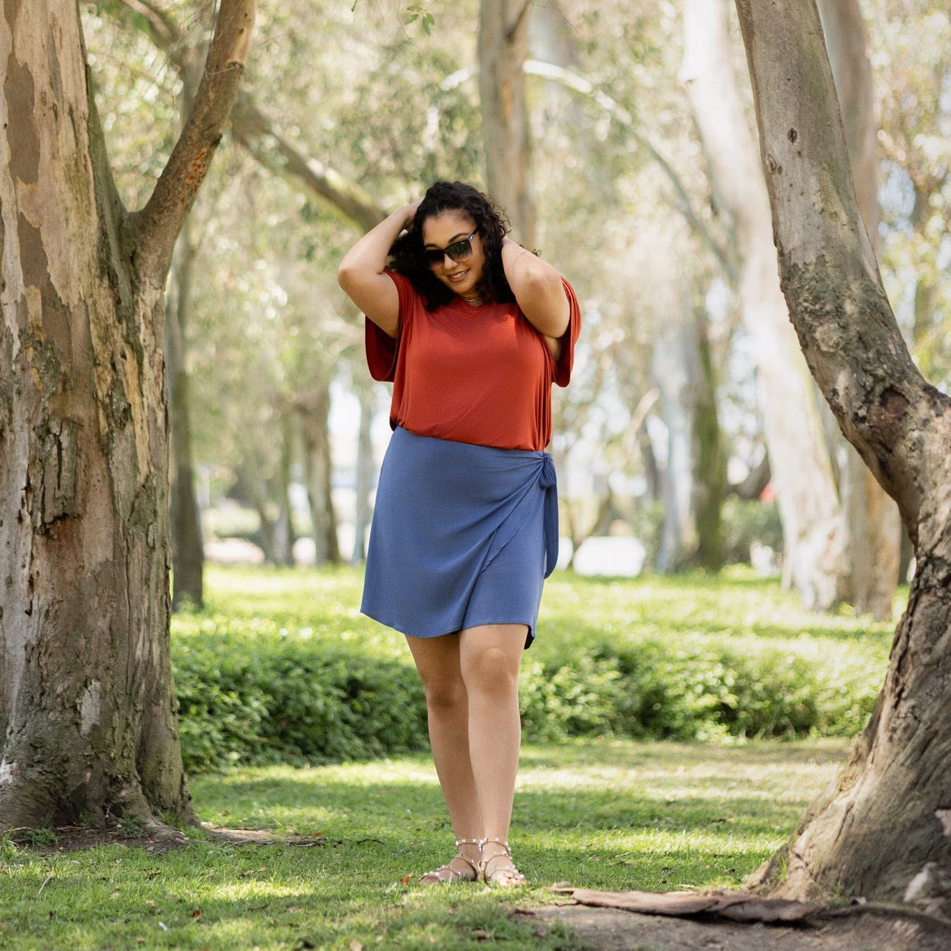Woman in an orange top and blue skirt standing in a park with trees and grass.