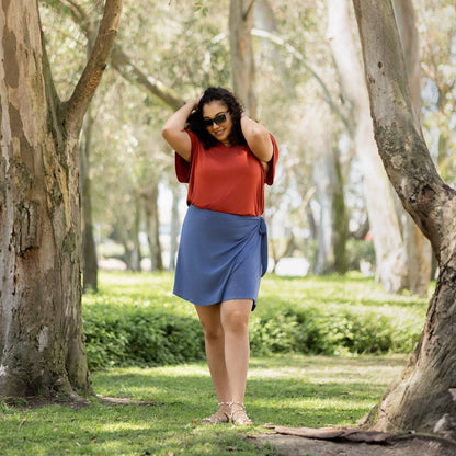 Woman in an orange top and blue skirt standing in a park with trees and grass.