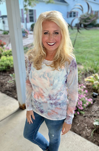 Woman wearing a tie-dye shirt and jeans standing outdoors with a house and garden in the background.